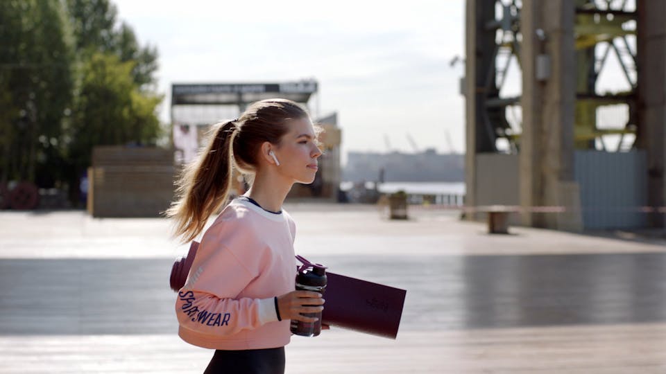 Woman Walking By The Seaport Holding A Yoga Mat And Drinking Water